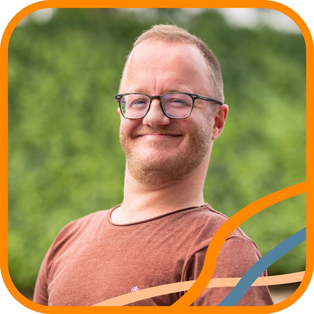 Portret Niels. Een witte man met kort rossig/blond haar kijkt lachend in de camera. Hij draagt een bril met blauw montuur en een terracotta-kleurig T-shirt.